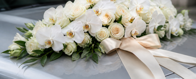 White orchid and cream rose wedding car bonnet arrangement tied with an ivory satin ribbon on a silver car